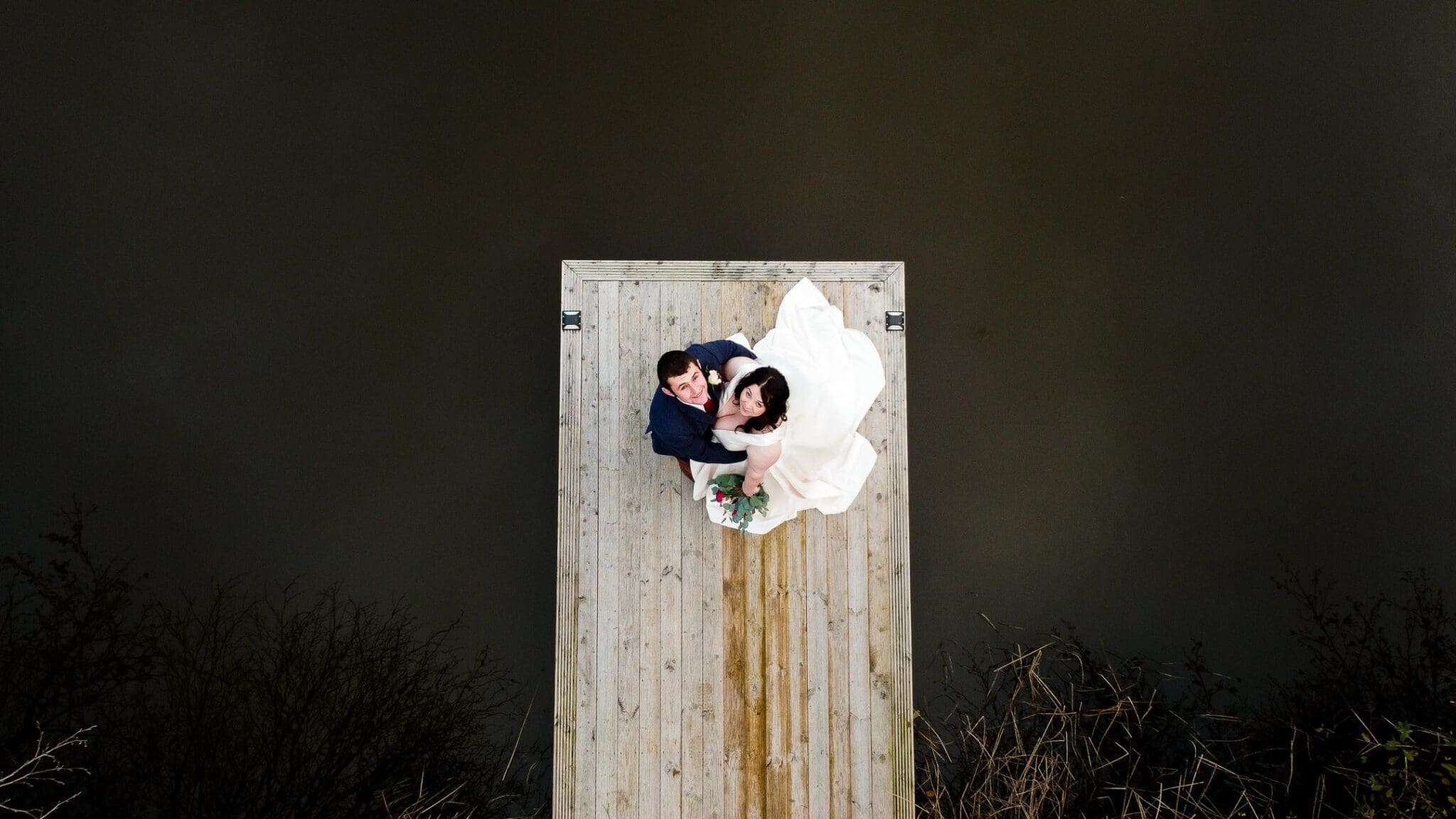 Bride & groom looking up on wooden jetty drone image 